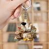 Keychain with a figurine of a girl reading a book on books, held by a hand in front of a blurred bookshelf.